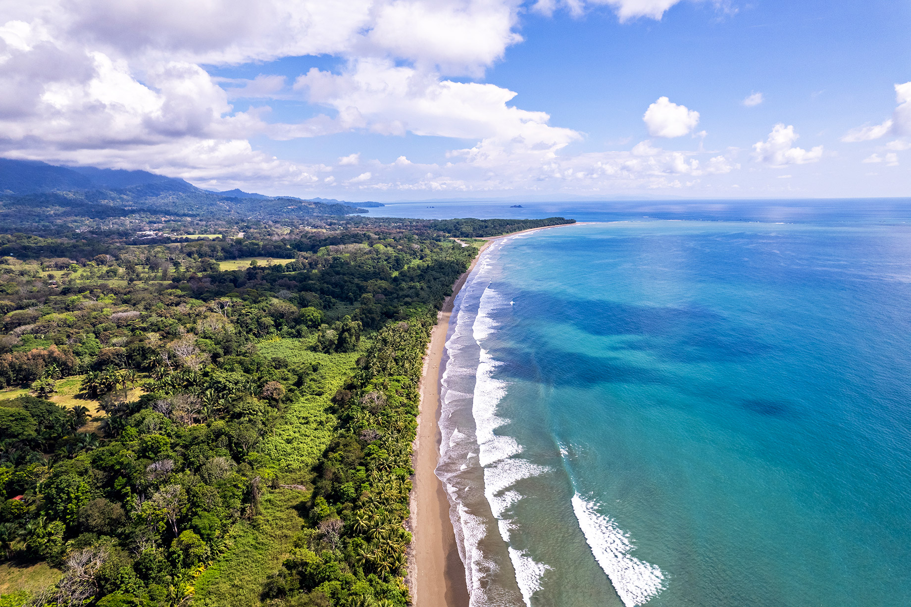 Drone image showing the lush jungle coastline and turquoise waters of Playa Hermosa stretching north toward Uvita’s Whale’s Tail, with rolling hills and tropical skies in the distance.