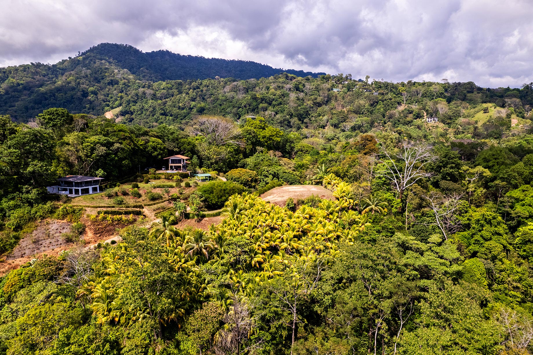 Drone image showcasing the terraced landscape of a coastal estate in Playa Hermosa, Uvita, featuring three existing residences surrounded by dense jungle, palm groves, and mountain backdrops.
