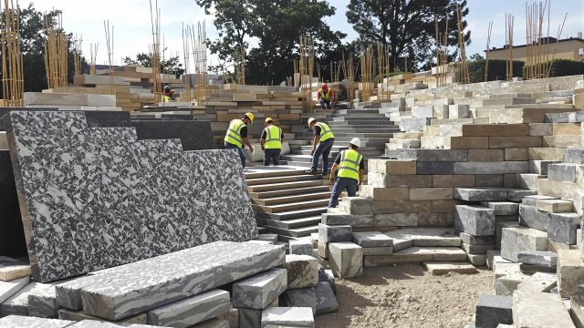 Construction site with various types of stones including granite, limestone, sandstone, fieldstone, slate, basalt, and marble, arranged in different formats and finishes. Workers are selecting and placing stones for different parts of a building and landscape project, showcasing the durability, natural beauty, and versatility of building with stone.