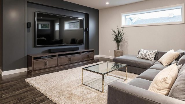 A luxury modern contemporary finished basement featuring a home theater setup with a large flat-screen TV, a comfortable seating area with a sofa and a coffee table, and an egress window. The space is designed to feel like an extension of the upstairs living area, with consistent decor and modern lighting.