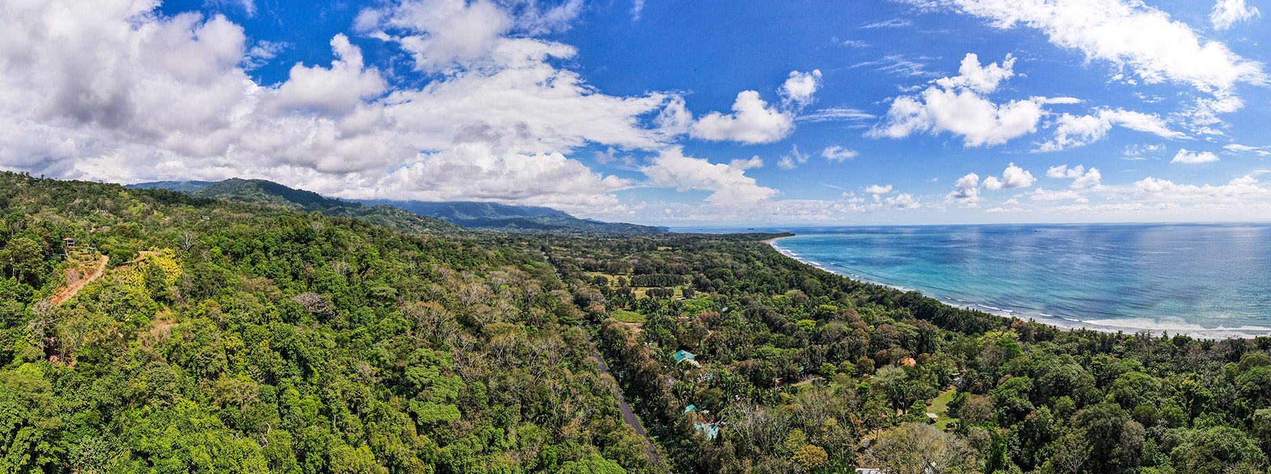 Aerial panorama of lush jungle ridgeline above Playa Hermosa with sweeping views of Costa Rica’s Whale’s Tail coastline and the Pacific Ocean under a vibrant blue sky.