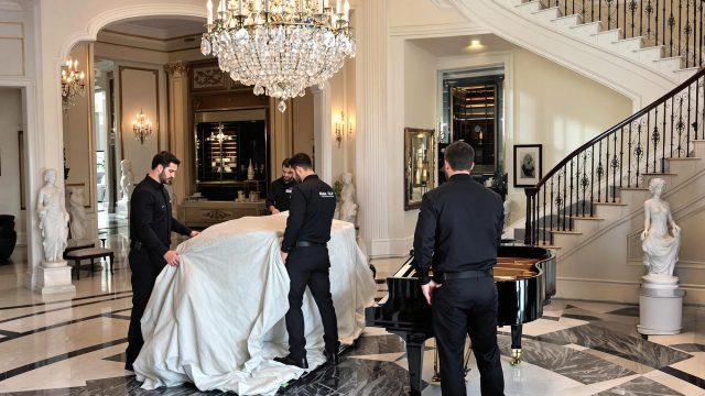 Three professional movers in black uniforms carefully covering a large piece of furniture with a protective cloth in a luxurious estate. The grand room features a chandelier, marble statues, a grand piano, and a spiral staircase, highlighting the sophistication and precision involved in luxury relocations.
