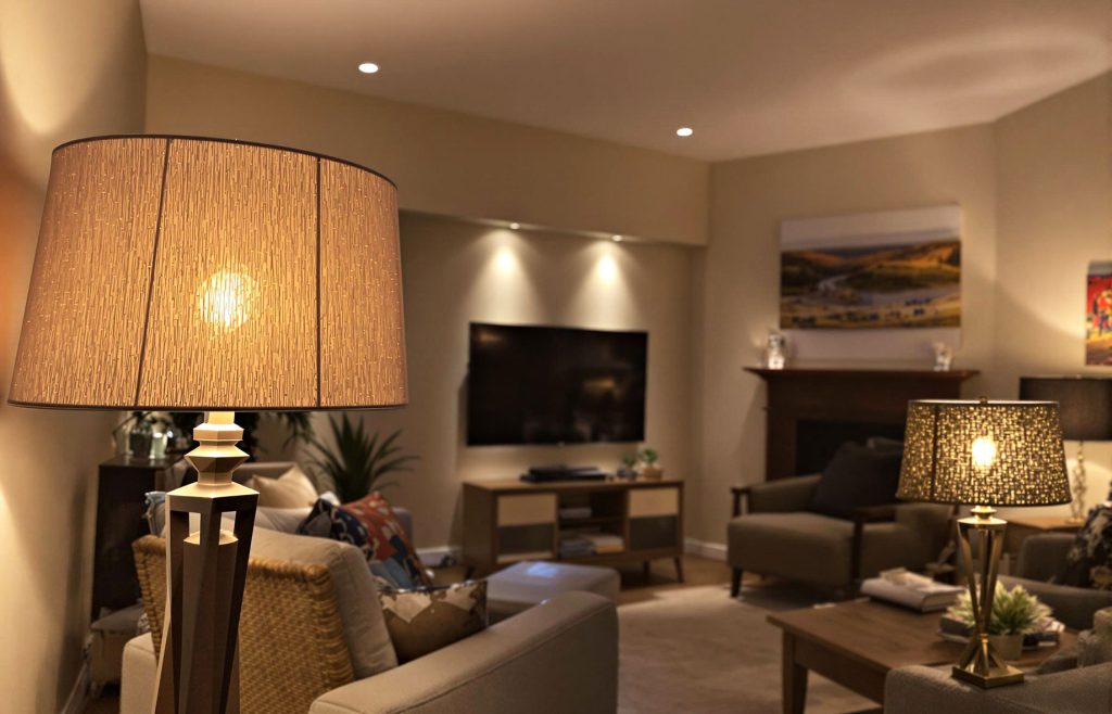 A cozy living room featuring layered lighting with a fabric-shaded floor lamp, recessed ceiling lights, a warm-toned table lamp, LED strips behind the television, and spotlights highlighting artwork, creating a warm and inviting atmosphere.