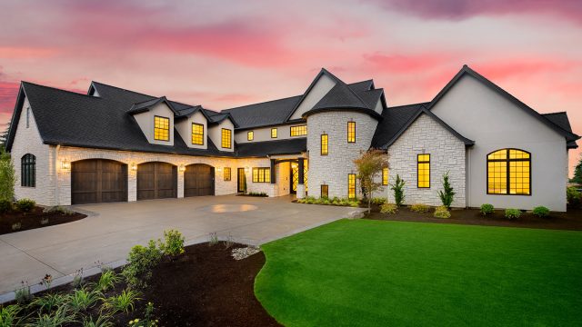 A stunning modern luxury estate with a mix of stone and stucco exterior, featuring multiple garage doors, large windows, and a beautifully landscaped front yard, all illuminated during a picturesque sunset.