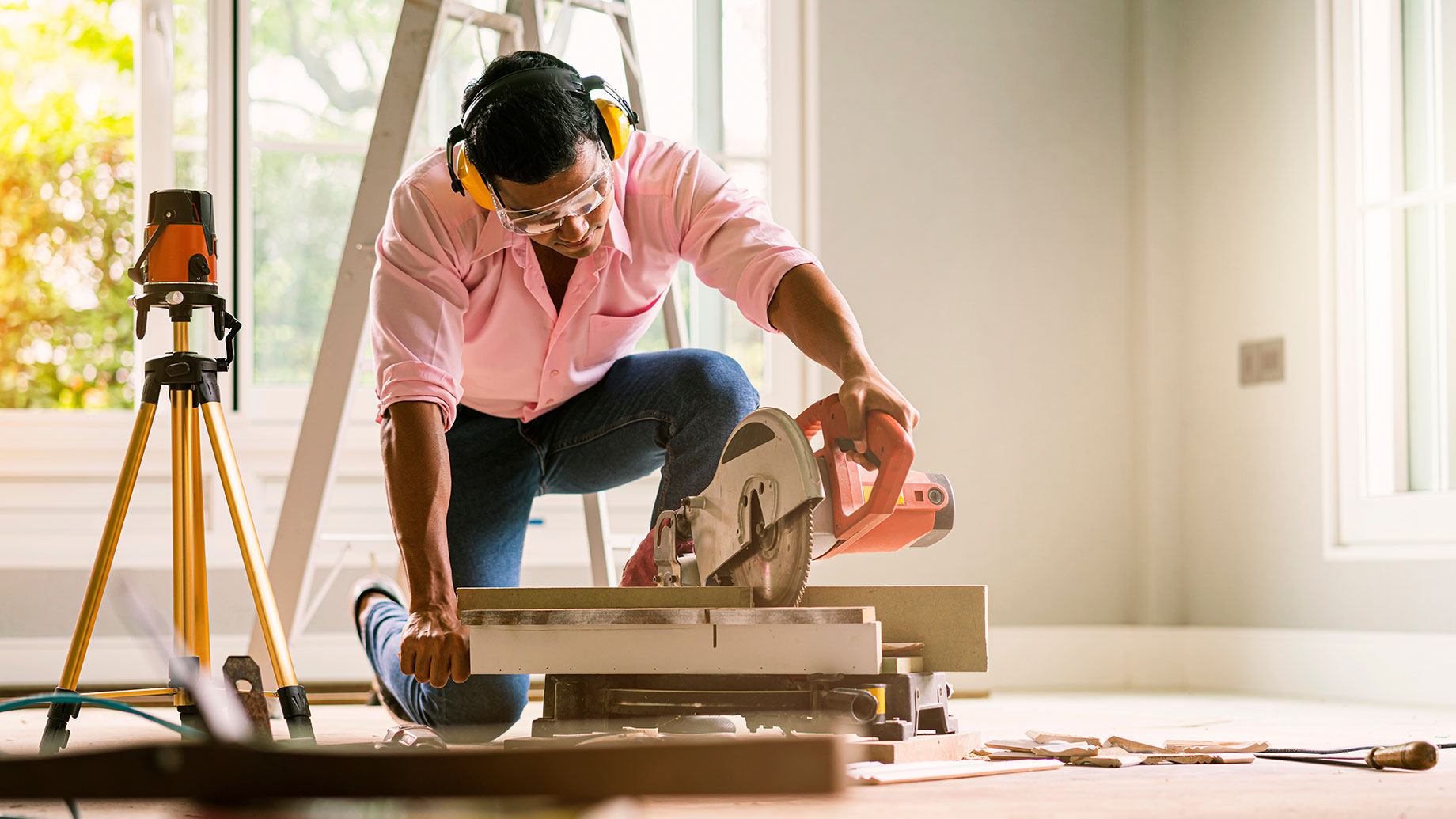 A construction worker wearing safety goggles and ear protection is using a circular saw to cut a piece of material during a home renovation project. A tripod-mounted laser level is visible in the background, indicating careful measurement and planning.