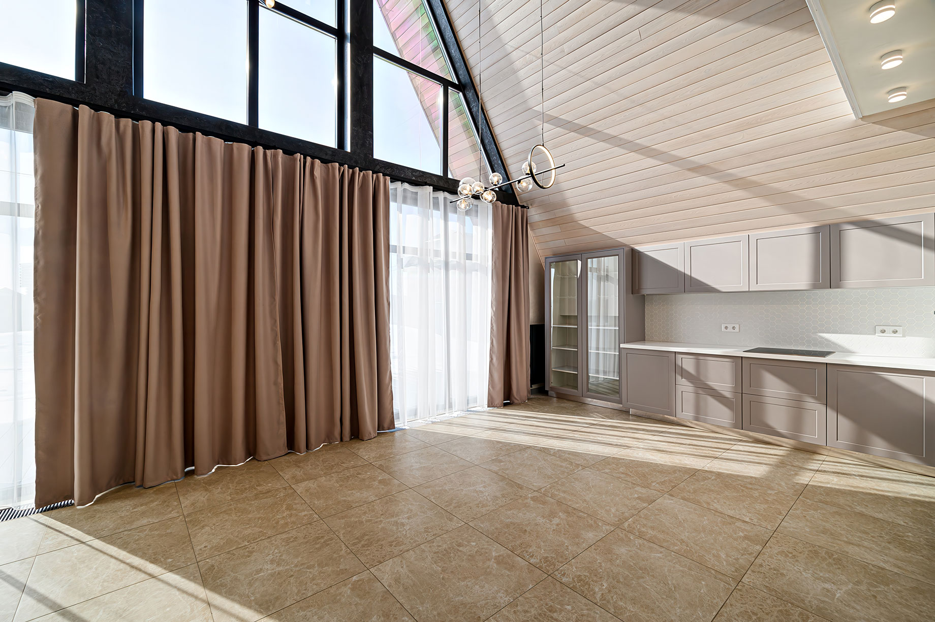Bright Interior with Tile Flooring, a Peaked Ceiling, Kitchen Cabinets, and Large Windows Covered with Neutral Curtains