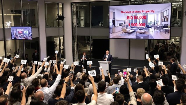 A lively auction room with potential buyers raising paddles and auctioneers engaging with the crowd, showcasing a unique property on a large screen and highlighting the competitive atmosphere.