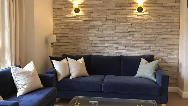 A cozy living room featuring a stone effect accent wall, a velvet sofa, linen curtains, and a glass coffee table, illuminated by wall sconces casting a warm glow.