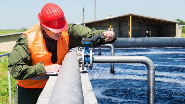 A technician wearing safety gear, including a red hard hat and an orange safety vest, inspects a valve on a large pipe at a wastewater treatment facility, ensuring the system's proper function and integrity.