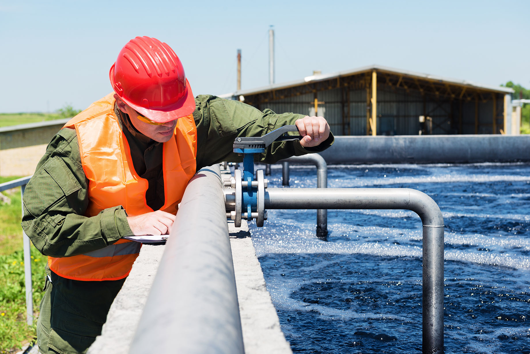 A technician wearing safety gear, including a red hard hat and an orange safety vest, inspects a valve on a large pipe at a wastewater treatment facility, ensuring the system's proper function and integrity.
