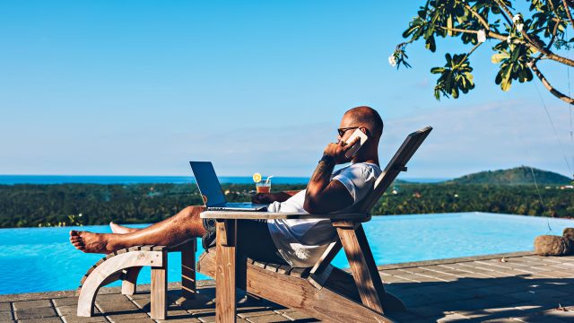 Young businessman resting on a sunbed, enjoying summertime at a resort beside a swimming pool, while working remotely on his laptop computer, connected to wireless internet and talking on his smartphone.