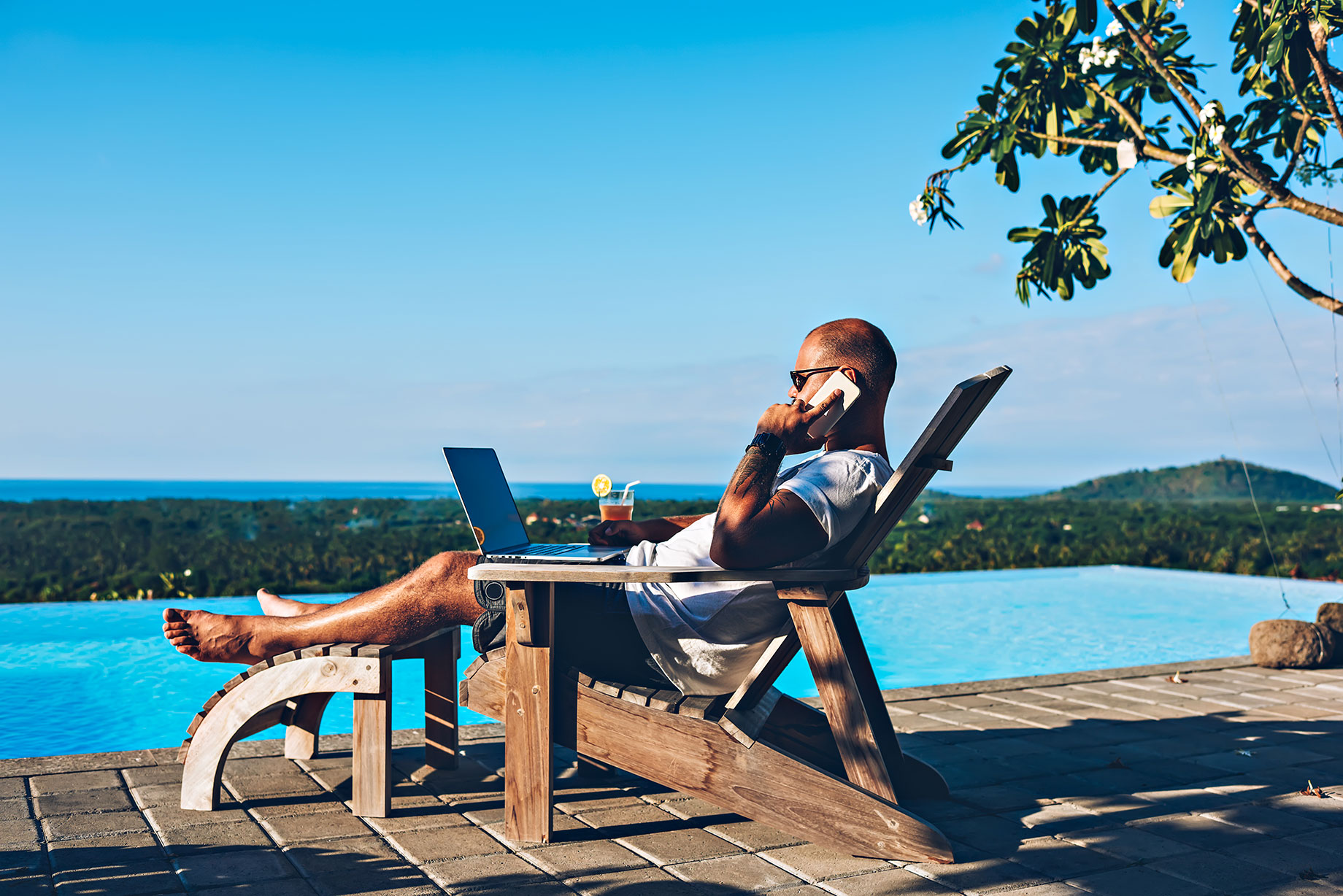 Young businessman resting on a sunbed, enjoying summertime at a resort beside a swimming pool, while working remotely on his laptop computer, connected to wireless internet and talking on his smartphone.