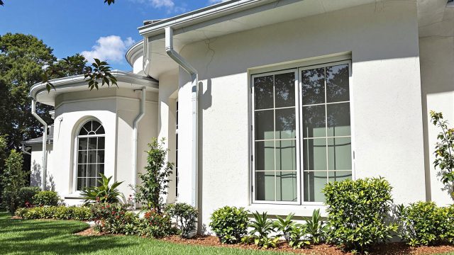A luxurious home exterior with a well-manicured lawn and elegant landscaping, showing subtle signs of potential issues such as small cracks in the walls, an uneven roofline, a clogged gutter, and a window with condensation.