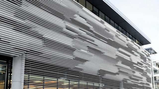 A modern building featuring aluminium batten cladding on its exterior façade. The linear aluminium profiles create dynamic shadow lines and visual texture, adding depth and architectural interest. The building is set in an urban environment, showcasing the material's versatility and aesthetic appeal in contemporary architecture.