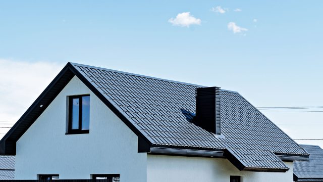 A modern house with a sleek, dark grey metal roof and a small attic window under a clear blue sky.