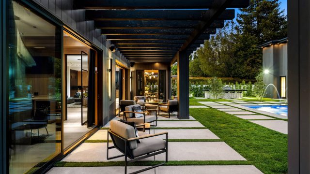 Modern luxury terrace at dusk with black pergola, lounge seating on lawn-inlaid pavers, pool with water jets, and sliding glass walls connecting to the interior.