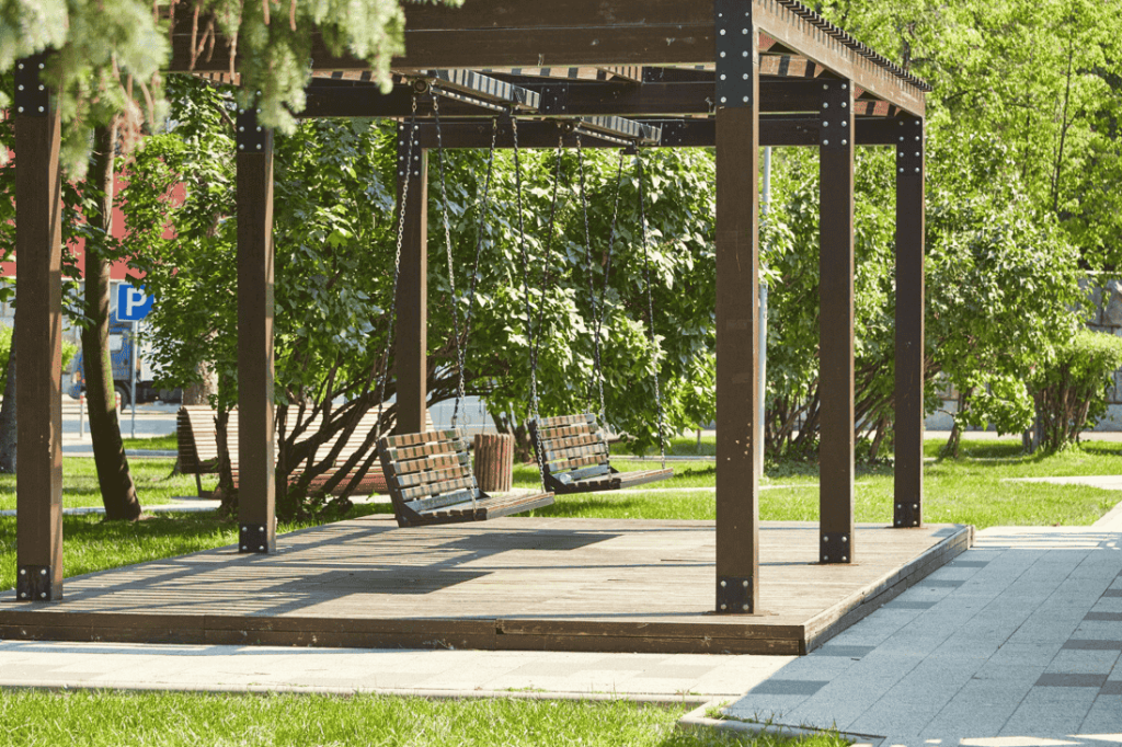 A shaded wooden pergola with two hanging swings in a landscaped green area, showcasing a relaxing outdoor feature in a luxury community.