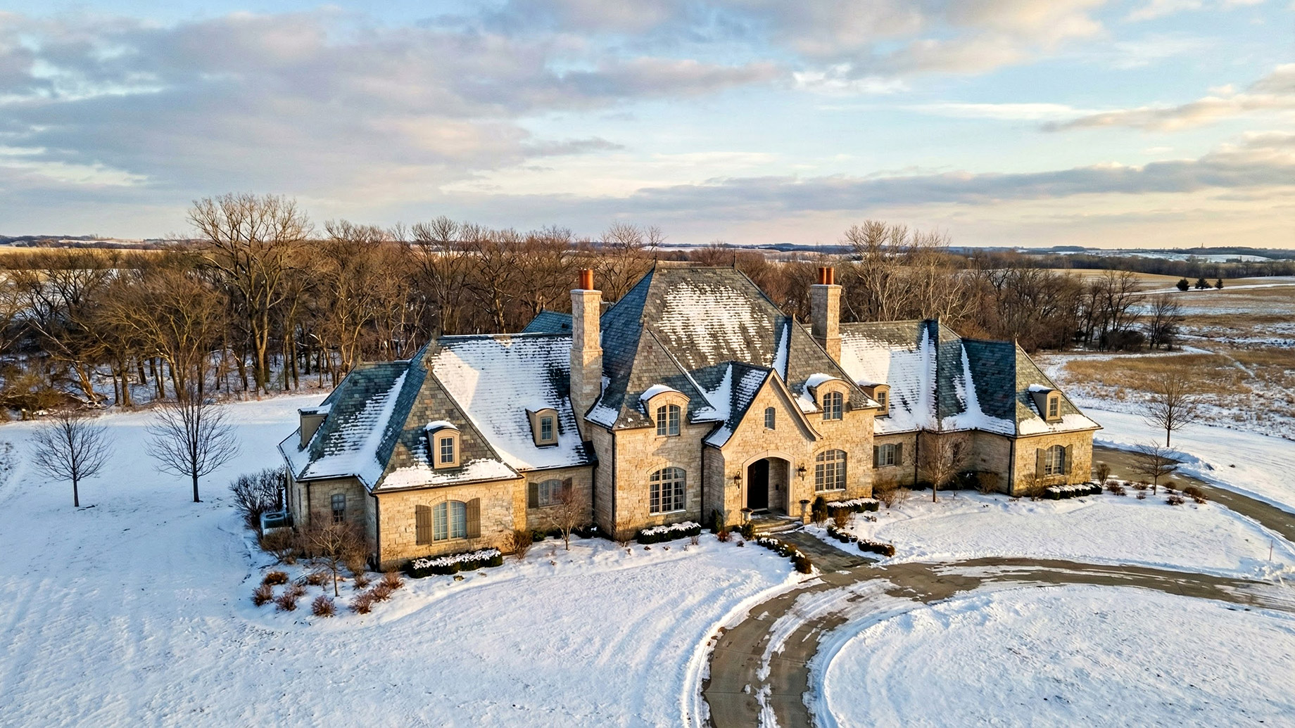 Aerial view of a sprawling French Provincial-style luxury estate with a prominent slate roof partially covered in snow, situated in a rural Midwest landscape with bare trees and snow-covered fields under a cloudy winter sky.