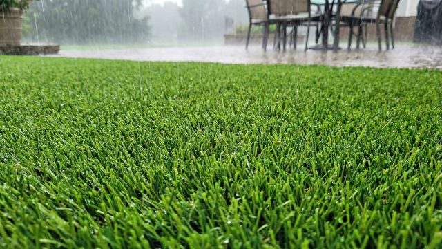 A close-up view of vibrant green artificial grass during a heavy rainstorm, demonstrating its ability to handle water without mud or puddling. Rain droplets are visible on the blades, while a wet patio area with outdoor furniture sits in the blurred background, highlighting the turf's resilience in extreme weather.