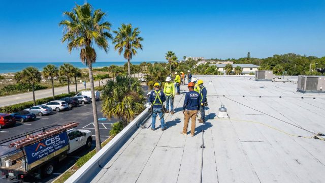 A professional crew from CES Commercial Roofing working on a flat white commercial roof in Palm Harbor, Florida, under a sunny blue sky with palm trees and the coast visible in the background.