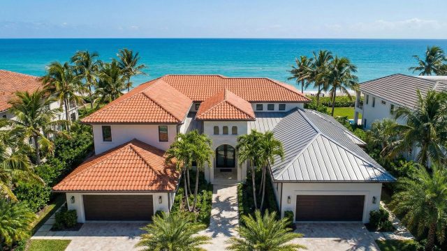 An aerial photograph of a sprawling luxury coastal residence in South Florida, featuring a combination roof with reddish-orange terra cotta tiles on the main structure and grey standing-seam metal roofing on the attached garage section, all framed by palm trees against a turquoise ocean background.