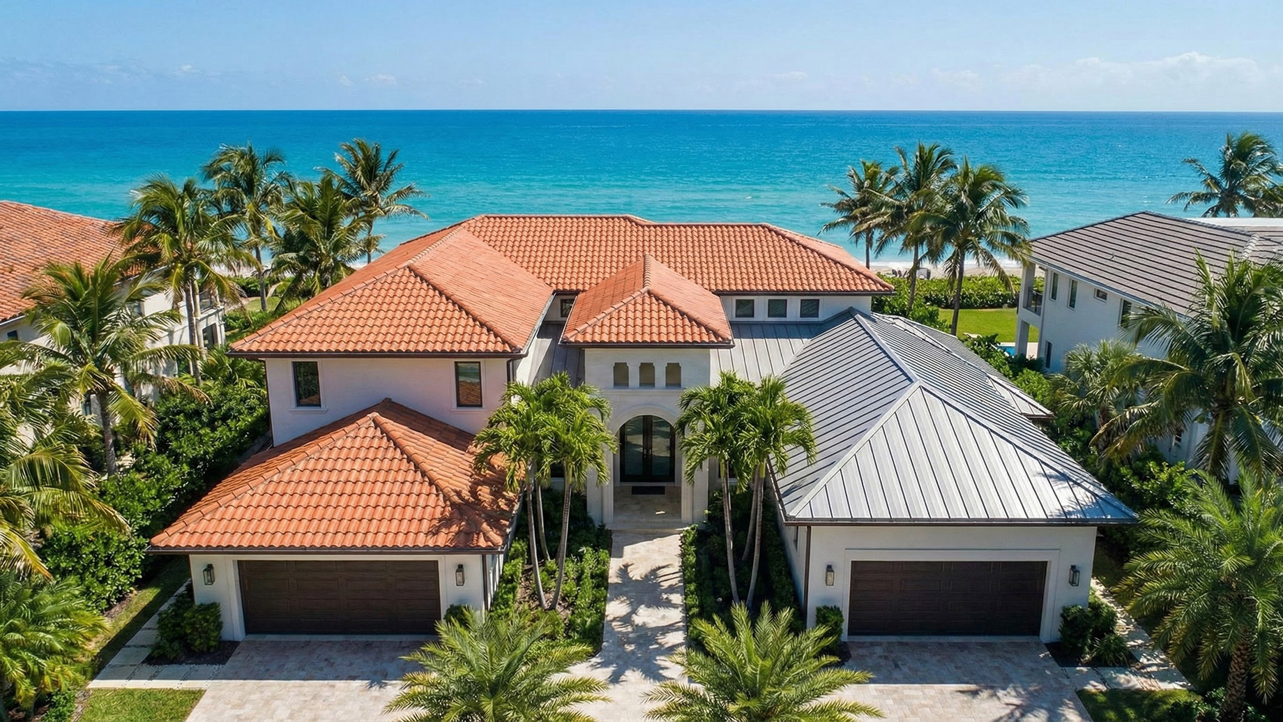 An aerial photograph of a sprawling luxury coastal residence in South Florida, featuring a combination roof with reddish-orange terra cotta tiles on the main structure and grey standing-seam metal roofing on the attached garage section, all framed by palm trees against a turquoise ocean background.