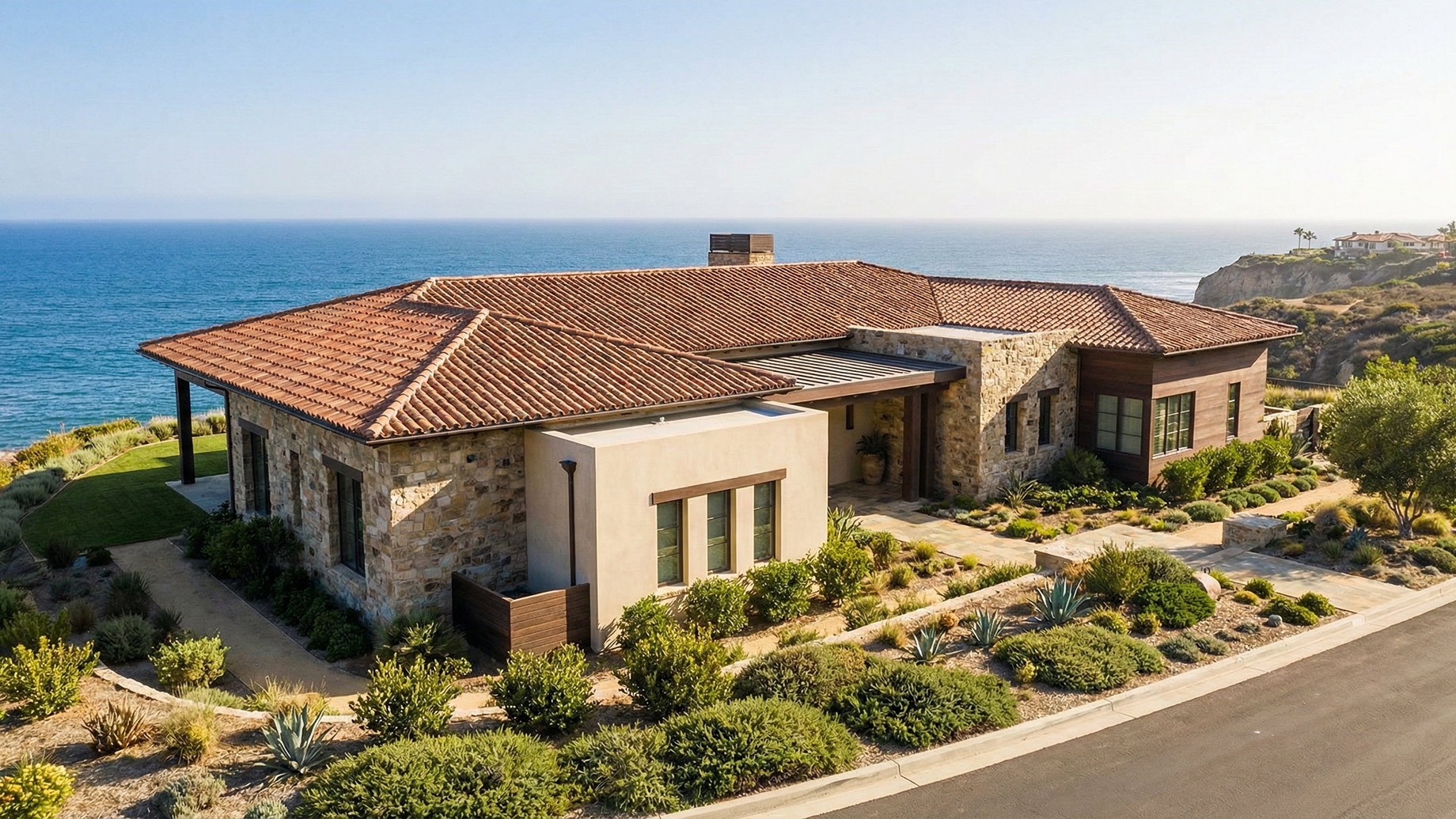 An aerial view of a luxurious, single-story home perched on a cliffside overlooking the blue ocean. The residence features a mix of weathered stone, beige stucco, and dark wood siding, topped with a prominent terracotta tiled roof. Landscaped gardens with drought-resistant plants surround the property, and a paved driveway leads to the entrance.