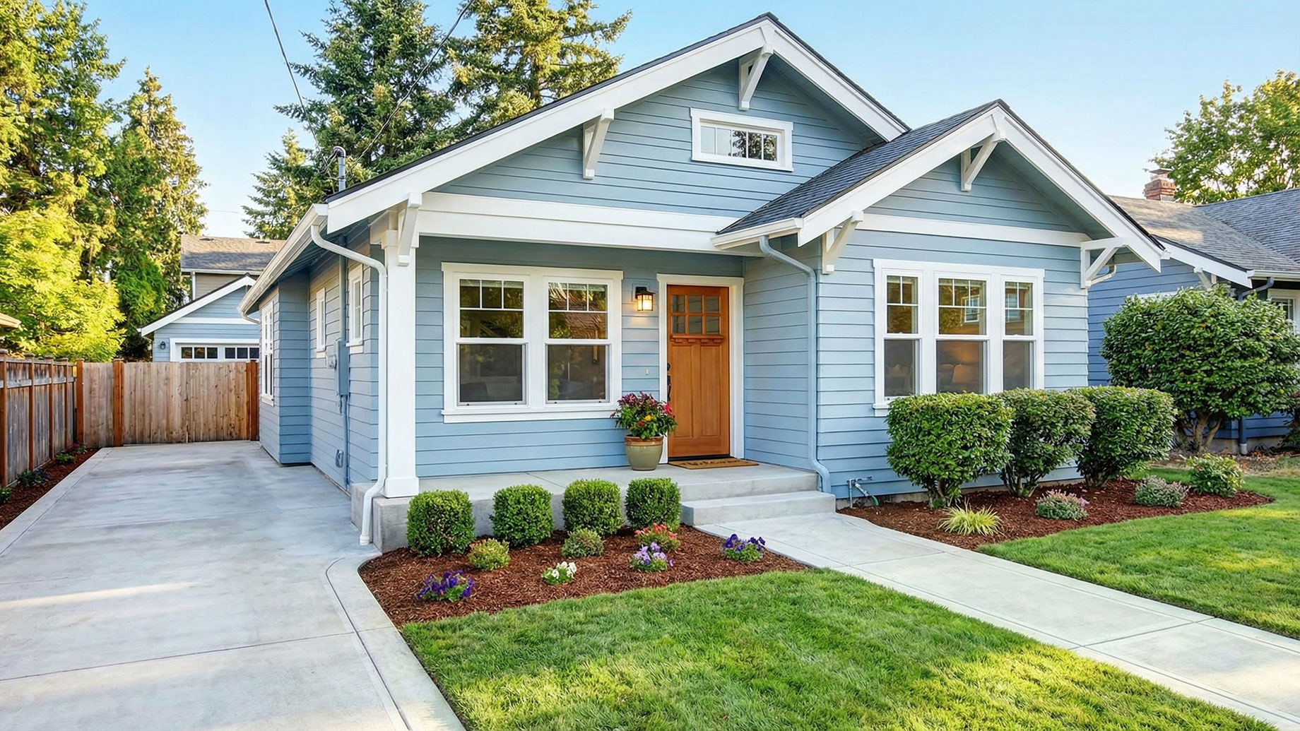 The exterior of a light blue Craftsman-style home featuring a pristine concrete driveway, freshly mulched garden beds, a manicured green lawn, and clean white trim, showcasing meticulous home maintenance.