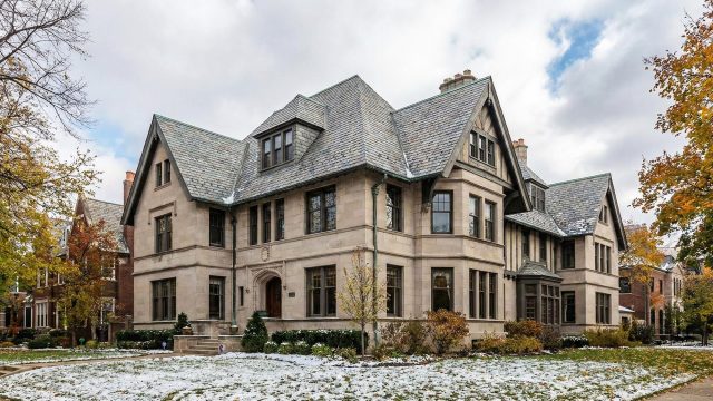 A wide-angle landscape photograph of a sprawling historic stone mansion in an established Chicago neighborhood, featuring an intricate, multi-gabled slate roof structure with a light dusting of snow on the shingles and surrounding landscaping during late autumn.
