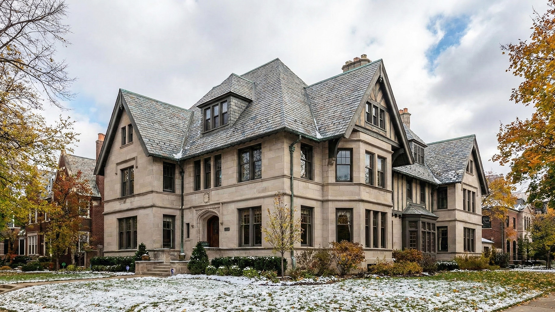 A wide-angle landscape photograph of a sprawling historic stone mansion in an established Chicago neighborhood, featuring an intricate, multi-gabled slate roof structure with a light dusting of snow on the shingles and surrounding landscaping during late autumn.