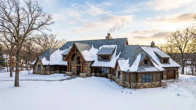 A large, luxurious stone and timber home in Minnesota is shown during a winter sunset. The complex, snow-covered roof features a mix of grey slate shingles and metal roofing on several dormers and turrets. Smoke rises from a central stone chimney. The grounds are blanketed in snow, and bare trees surround the estate.