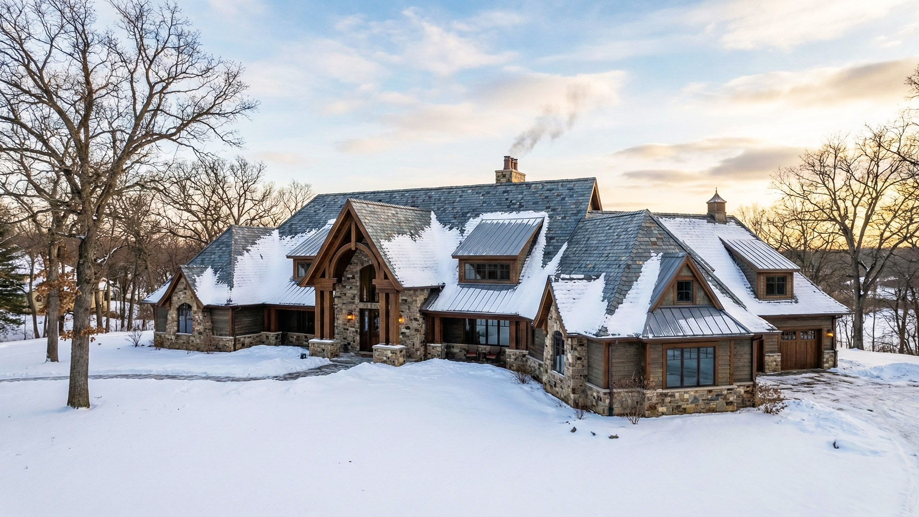 A large, luxurious stone and timber home in Minnesota is shown during a winter sunset. The complex, snow-covered roof features a mix of grey slate shingles and metal roofing on several dormers and turrets. Smoke rises from a central stone chimney. The grounds are blanketed in snow, and bare trees surround the estate.