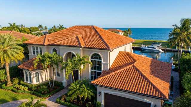 Aerial view of a sprawling luxury home in Cutler Bay, Florida, featuring a pristine terracotta clay tile roof, a private boat dock, and lush tropical landscaping set against a backdrop of blue ocean waters.