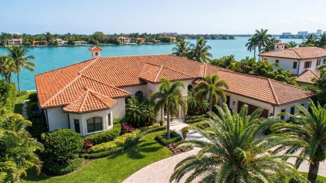 Aerial view of a luxury waterfront estate in Miami featuring a pristine terracotta clay tile roof and lush tropical landscaping, highlighting the importance of proper roof maintenance in South Florida to protect against coastal weather conditions.