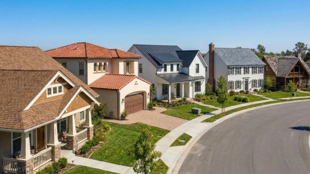 A panoramic view showing a row of homes with different architectural styles, each featuring a distinct roofing material—including terracotta clay tiles, standing seam metal, asphalt shingles, gray slate, and wood shakes—demonstrating how specific roofs complement different house designs.