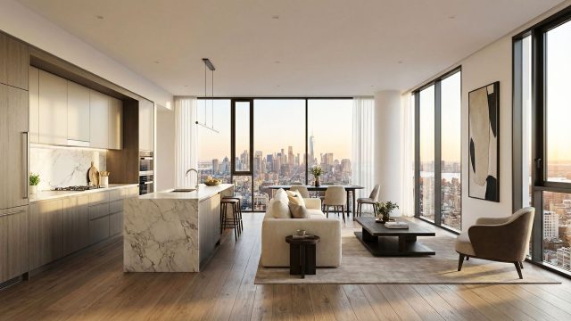 Interior of a spacious, open-concept luxury apartment featuring a modern kitchen with a marble island and a living area with contemporary beige furniture. Floor-to-ceiling windows run the length of the room, offering a sweeping city skyline view bathed in warm sunset light.