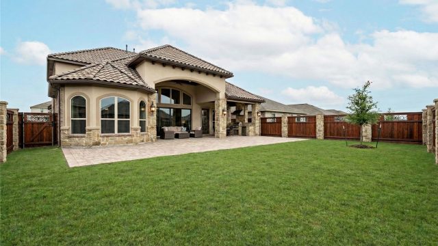 A wide, high-angle shot of a large, empty backyard of a luxury modern home featuring a manicured flat green lawn, a high-end horizontal wood slat privacy fence, and expansive glass windows, illustrating a space that feels vacant despite its size.