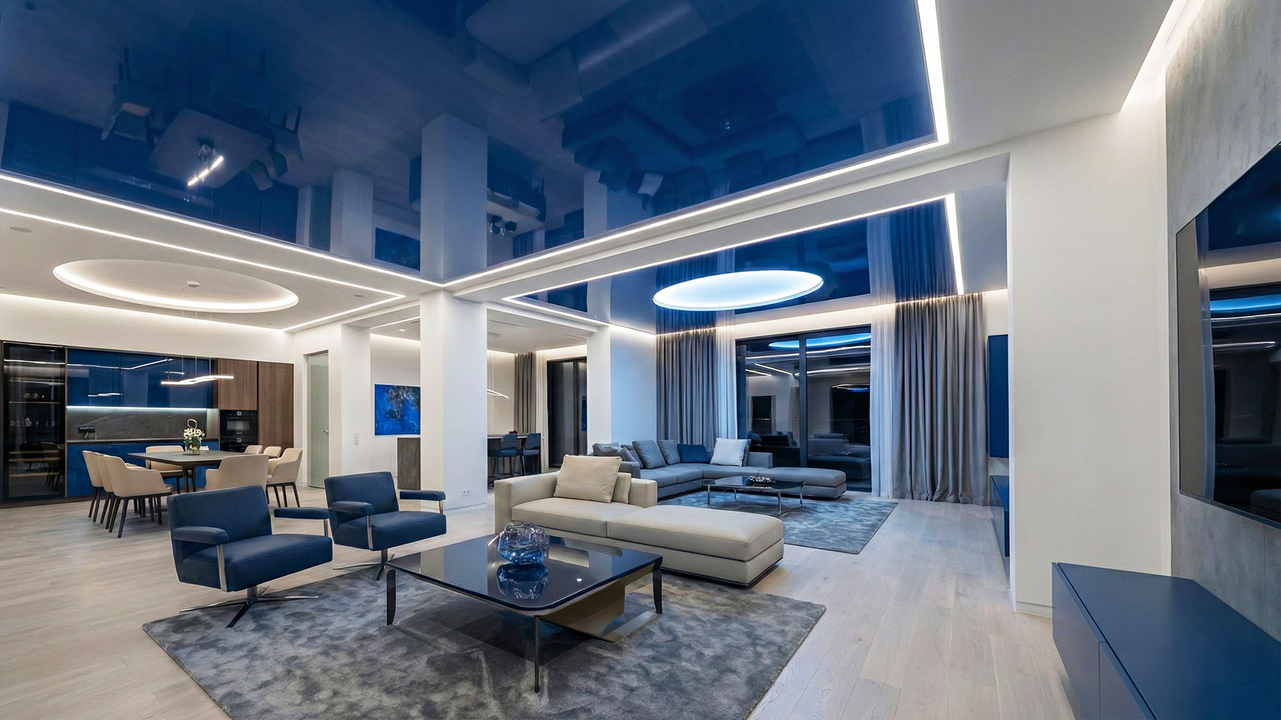 A spacious contemporary living room showcasing a seamless, high-gloss stretch ceiling with integrated LED lighting; the reflective surface mirrors the interior decor to create a sense of depth, height, and modern elegance.