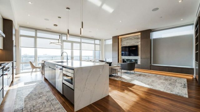 A spacious modern luxury home interior featuring an open-concept layout with a large white marble kitchen island, sleek cabinetry, hardwood floors, and floor-to-ceiling windows filling the room with natural light.