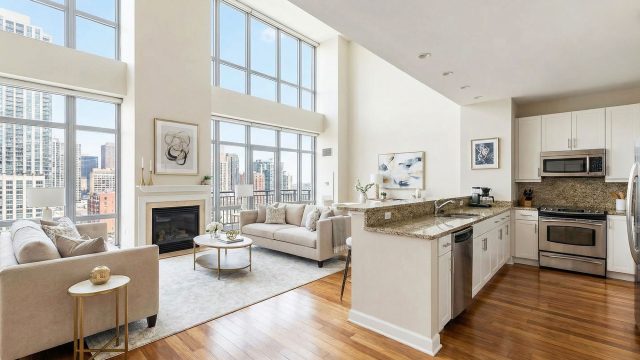 A spacious, modern luxury apartment interior featuring an open-concept living room and kitchen with high ceilings, floor-to-ceiling windows revealing a city skyline, hardwood flooring, granite countertops, and stainless steel appliances.