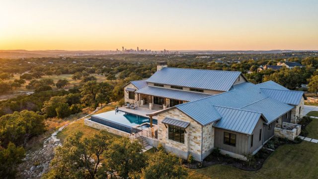 A sprawling luxury home in the Texas Hill Country featuring a prominent standing seam metal roof, stone architecture, and an infinity pool, overlooking the distant Austin city skyline during a golden sunset.