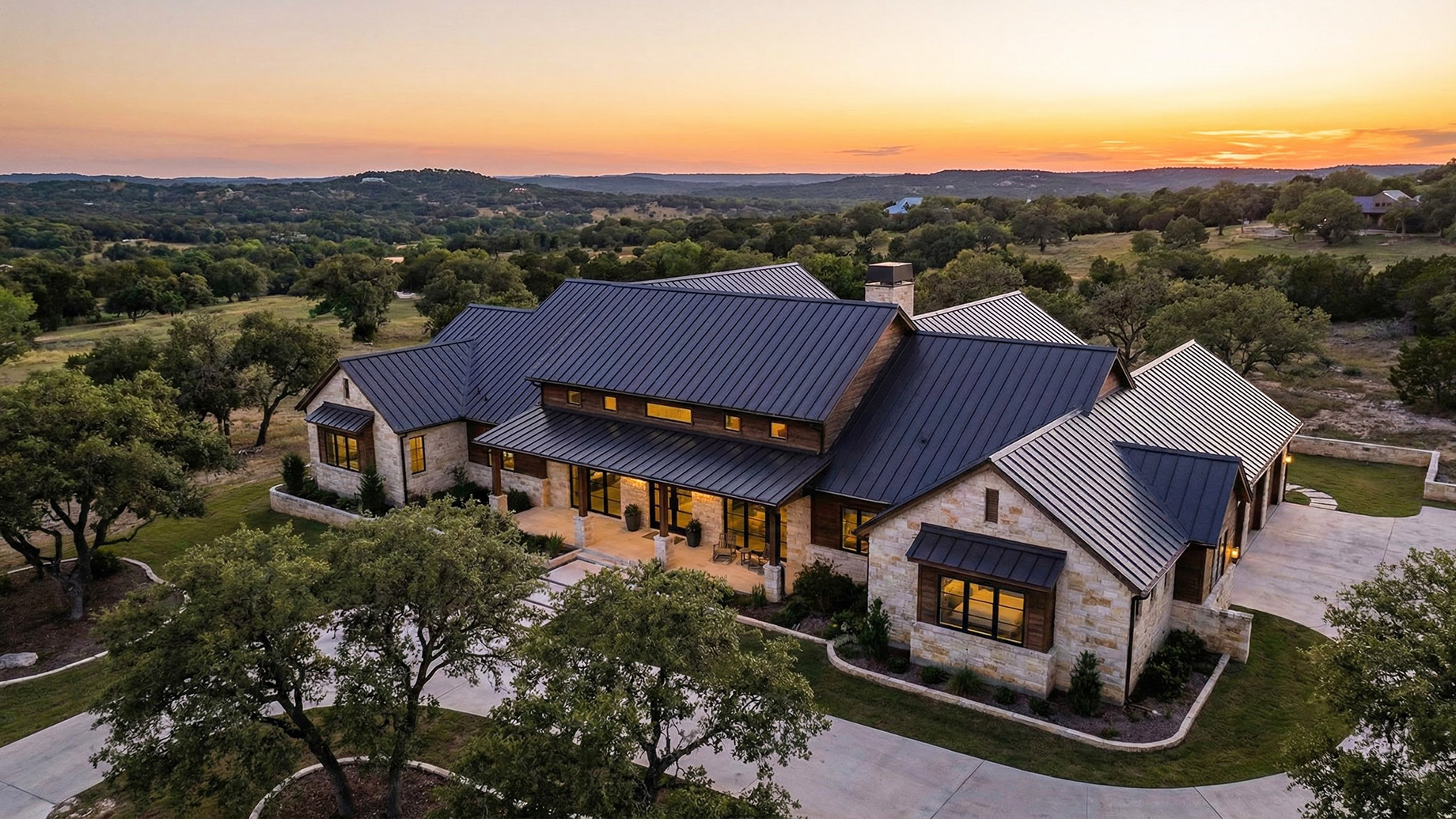 An aerial view of a sprawling luxury residence in the Texas Hill Country at sunset, featuring a prominent dark standing-seam metal roof and stone architecture surrounded by oak trees and rolling hills.