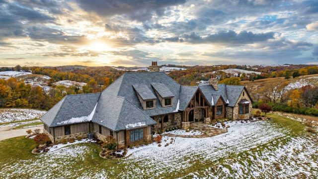 An aerial photograph of a sprawling luxury estate located in the rolling hills of the Midwest. The home features a large, high-quality slate roof, stone and timber siding, and is lightly dusted with an early winter snow under a dramatic sunset sky.