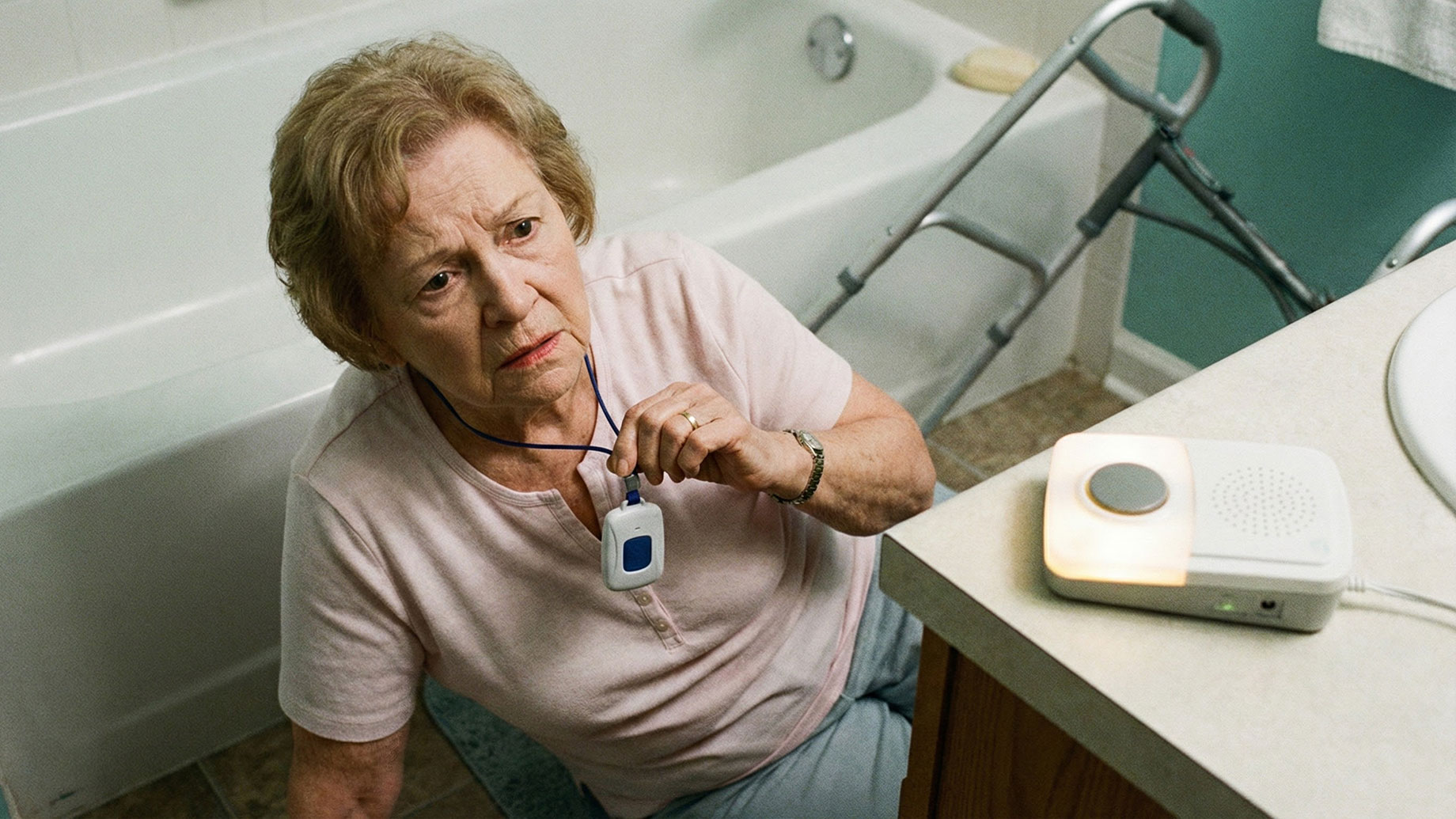 An elderly woman, who appears to have fallen, sits on a bathroom floor and holds her medical alert pendant, with a walker and base station visible.