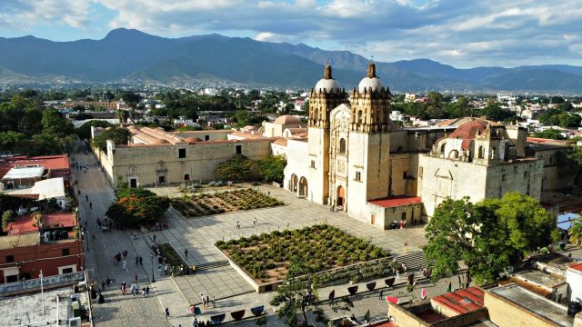 A high-angle, wide shot of the Templo de Santo Domingo de Guzmán in Oaxaca City. The massive, sand-colored stone colonial complex features two prominent bell towers with tiled domes and an expansive stone plaza in the foreground. Lush green mountains rise in the background under a blue sky with scattered white clouds. To the left, a pedestrian street is lined with colonial buildings, and small clusters of people are scattered across the courtyard.