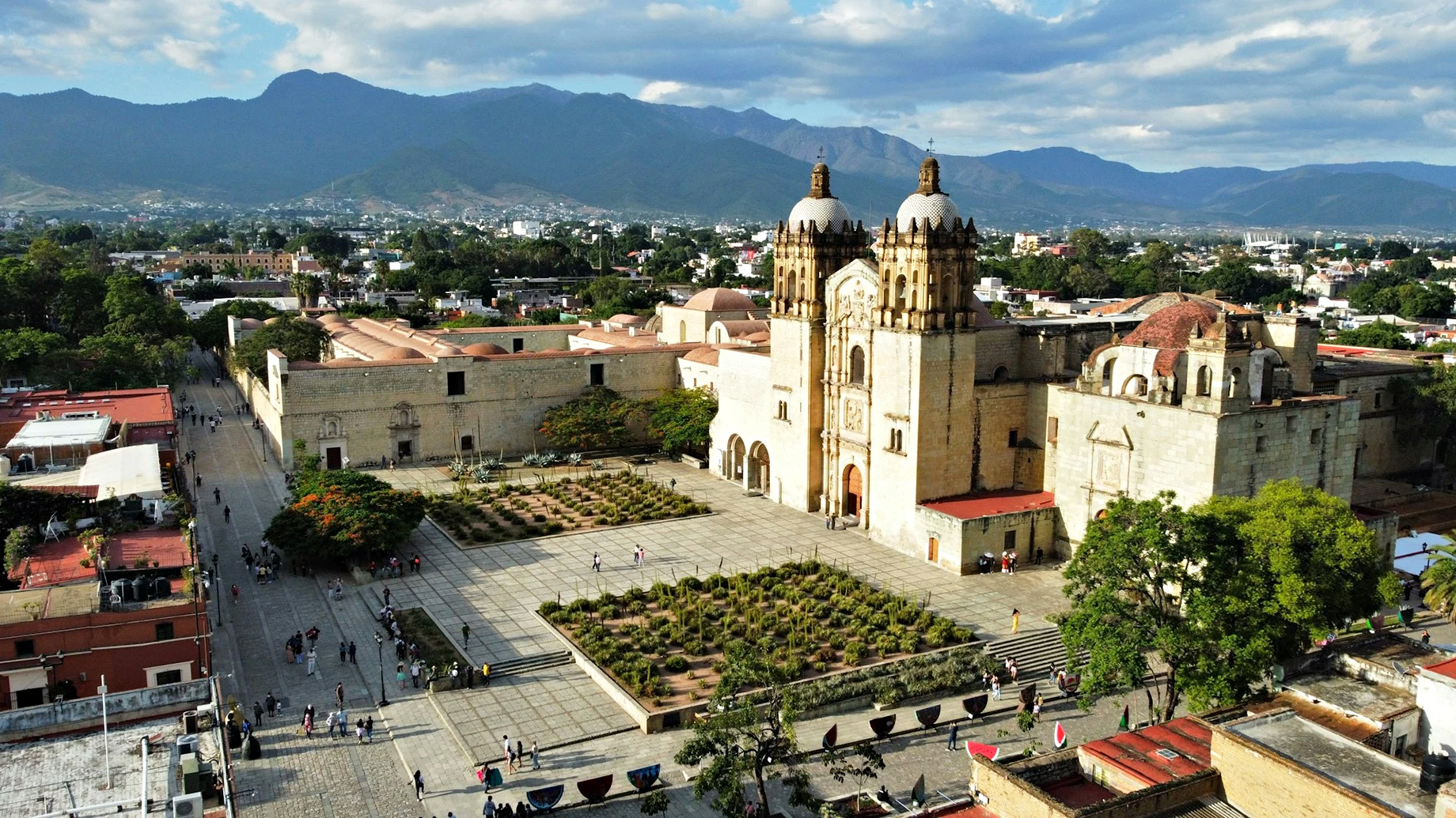 A high-angle, wide shot of the Templo de Santo Domingo de Guzmán in Oaxaca City. The massive, sand-colored stone colonial complex features two prominent bell towers with tiled domes and an expansive stone plaza in the foreground. Lush green mountains rise in the background under a blue sky with scattered white clouds. To the left, a pedestrian street is lined with colonial buildings, and small clusters of people are scattered across the courtyard.