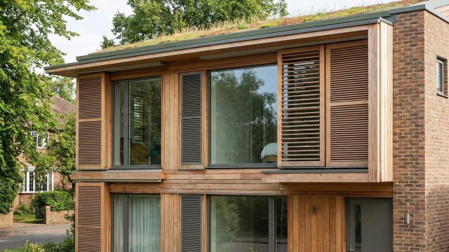 A modern two-story home in a leafy UK setting, featuring timber cladding, brickwork, and large windows with prominent external wooden louvered shutters (brise-soleil) for solar shading. A green sedum roof tops the structure, illustrating key passive design strategies.