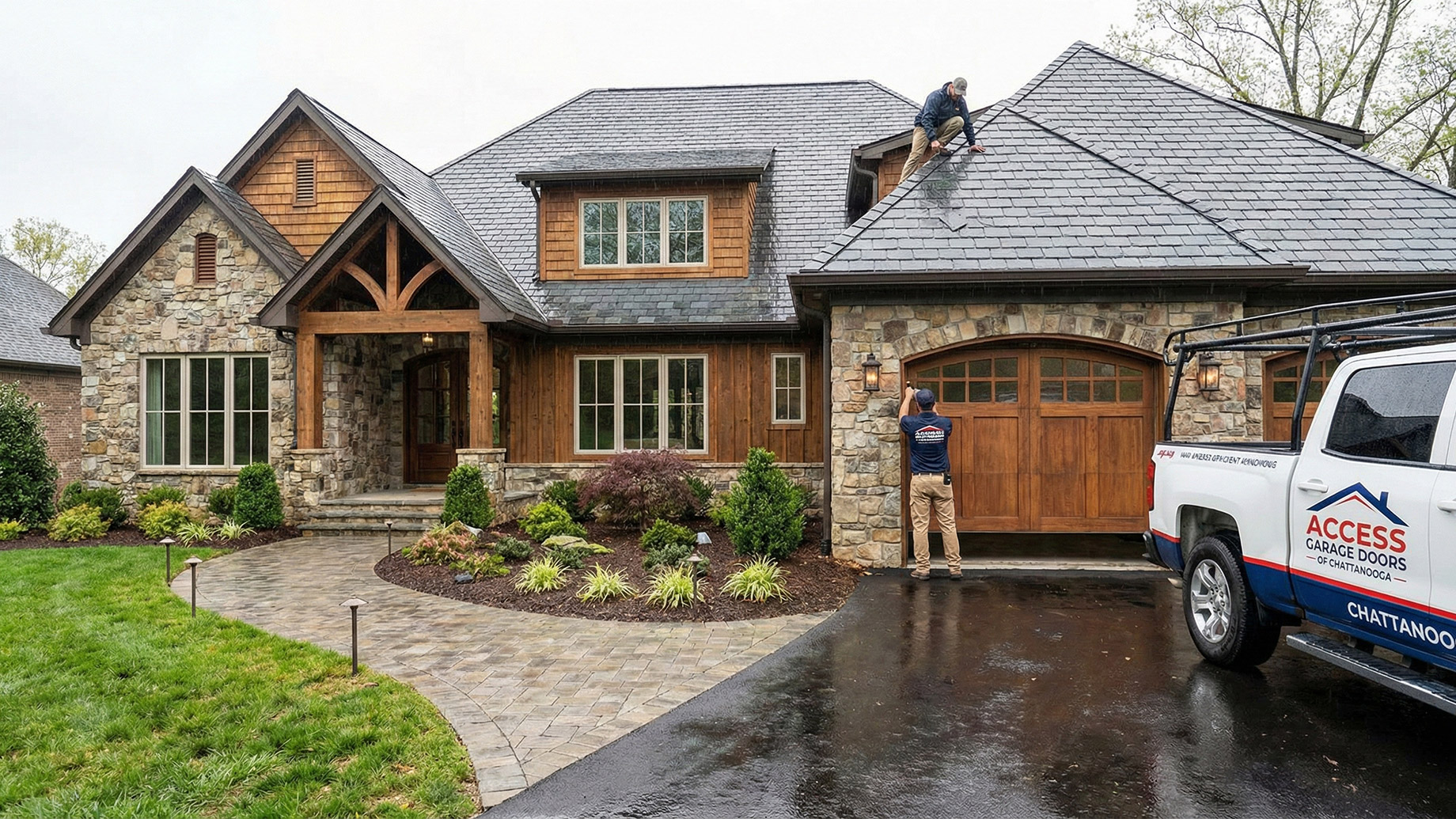 A luxury stone and wood home with a wet driveway, featuring a worker inspecting the slate roof and another technician next to a custom wooden garage door with an "Access Garage Doors" truck parked nearby.