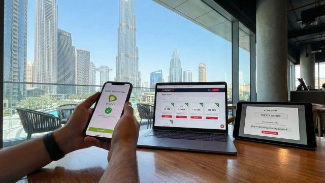 A workspace featuring a smartphone displaying the Etisalat logo, a laptop showing the Paynet.red online recharge interface with various AED voucher denominations (30, 55, 110, 210), and a tablet displaying a digital e-voucher code, all set against a bright window view of the Dubai skyline and Burj Khalifa.