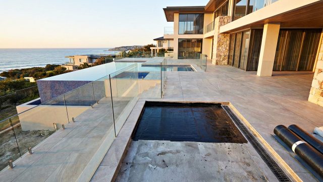 A section of concrete substrate on a luxury coastal home's multi-level patio is covered with a black liquid waterproofing membrane, next to rolls of membrane material and a linear drainage channel. The finished large-format tiled area with glass balustrades, an infinity pool, and the ocean view at sunset are visible in the background.